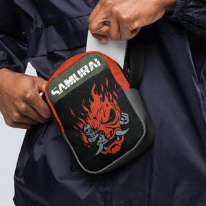 Könnte beinhalten: Eine kleine Umhängetasche in Schwarz und Rot mit einer Grafik eines stilisierten Robotergesichts, das von Flammen umgeben ist. Das Wort SAMURAI ist in weißen Großbuchstaben oben auf der Tasche aufgedruckt, die einen verstellbaren Riemen hat.