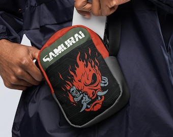 May include: A small crossbody bag featuring a black and red color scheme with a graphic of a stylized robotic face surrounded by flames. The word SAMURAI is printed in white capital letters at the top of the bag, which has an adjustable strap.