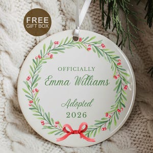 May include: A round white ceramic ornament featuring a green wreath with red berries and a red bow at the bottom. The text reads: OFFICIALLY Emma Williams Adopted 2026. The ornament hangs from a white ribbon and is designed for holiday decoration.