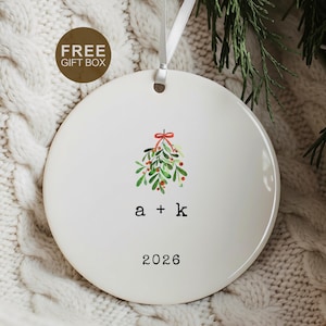 May include: A round white ceramic ornament featuring a watercolor illustration of green mistletoe with a red bow. Below the mistletoe, the lowercase letters a + k are printed in black, followed by the year 2026. A white ribbon is attached to the top.