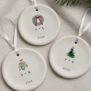 May include: Three round white ceramic ornaments with white ribbon hangers. Each features a watercolor illustration: a wreath with a red bow, a mistletoe sprig, or a pine tree. Below each graphic are lowercase initials and the year 2026.