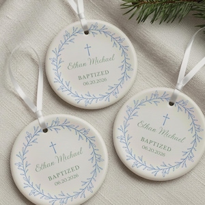 May include: Three round white ceramic ornaments with white satin ribbons. Each features a blue printed wreath, a small blue cross, and the text Ethan Michael, BAPTIZED, and 06.20.2026. The design is centered on the smooth, flat surface of each ornament.