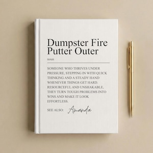 May include: A white hardcover journal with black text that reads Dumpster Fire Putter Outer, noun, followed by a definition describing someone who thrives under pressure, and the text See Also: Amanda, accompanied by a gold metal ballpoint pen.