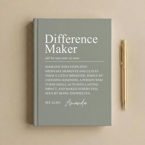May include: A sage green hardcover journal featuring the white printed text Difference Maker, a dictionary-style definition, and the name Amanda, accompanied by a gold-toned metal ballpoint pen positioned vertically to the right of the book.