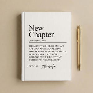 May include: A white hardcover journal featuring the black printed text New Chapter, a dictionary-style definition, and the name Amanda in script. A gold metal pen is positioned vertically to the right of the journal on a neutral background.