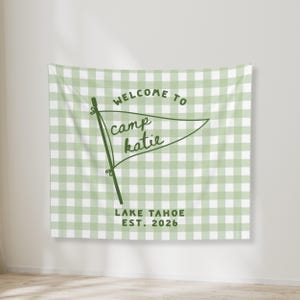 May include: A rectangular wall tapestry featuring a light sage green and white gingham check pattern. Centered is a dark green pennant flag graphic with the text "WELCOME TO camp katie" and "LAKE TAHOE EST. 2026" printed in dark green lettering.