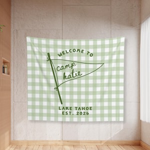 May include: A rectangular wall tapestry featuring a light sage green and white gingham check pattern. Centered is a dark green pennant flag graphic with the text "WELCOME TO camp katie" and "LAKE TAHOE EST. 2026" printed in dark green lettering.