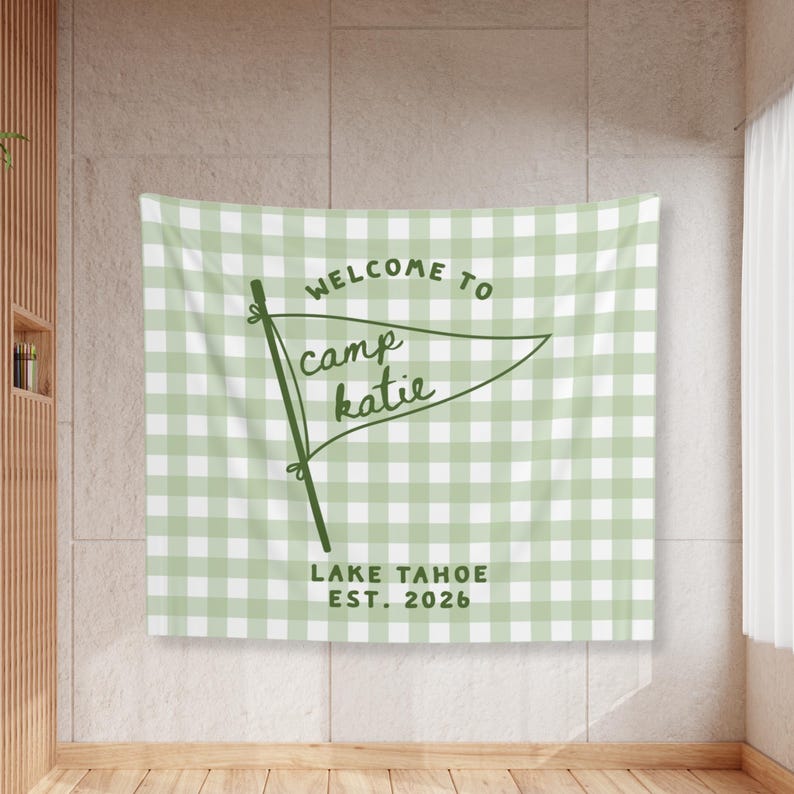 May include: A rectangular wall tapestry featuring a light sage green and white gingham check pattern. Centered is a dark green pennant flag graphic with the text "WELCOME TO camp katie" and "LAKE TAHOE EST. 2026" printed in dark green lettering.