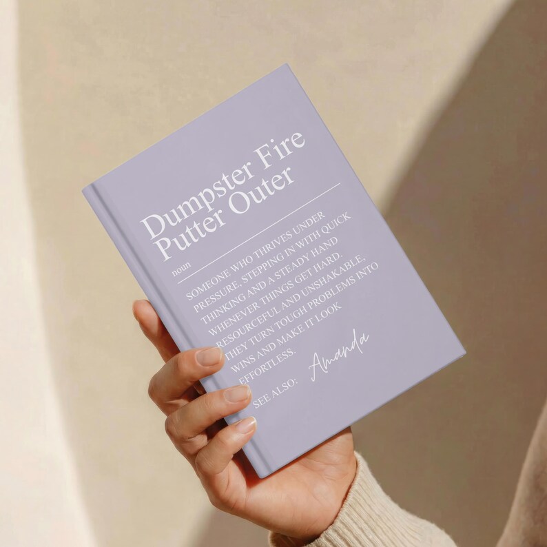 May include: A light purple hardcover journal featuring the white printed text Dumpster Fire Putter Outer, followed by a dictionary-style definition and the name Amanda. The book cover has a matte finish and displays a clean, modern layout.