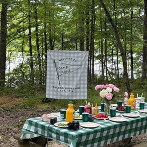 May include: A rectangular white and light green gingham fabric banner hanging outdoors, featuring the text "WELCOME TO camp rosie" with a pennant graphic and "POCONO MOUNTAINS EST. 1996" printed in dark green ink on the front surface.