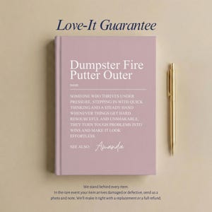 May include: A dusty rose hardcover journal featuring the white printed text Dumpster Fire Putter Outer, a definition of the term, and the name Amanda, accompanied by a gold-toned metal pen positioned vertically to the right of the book.