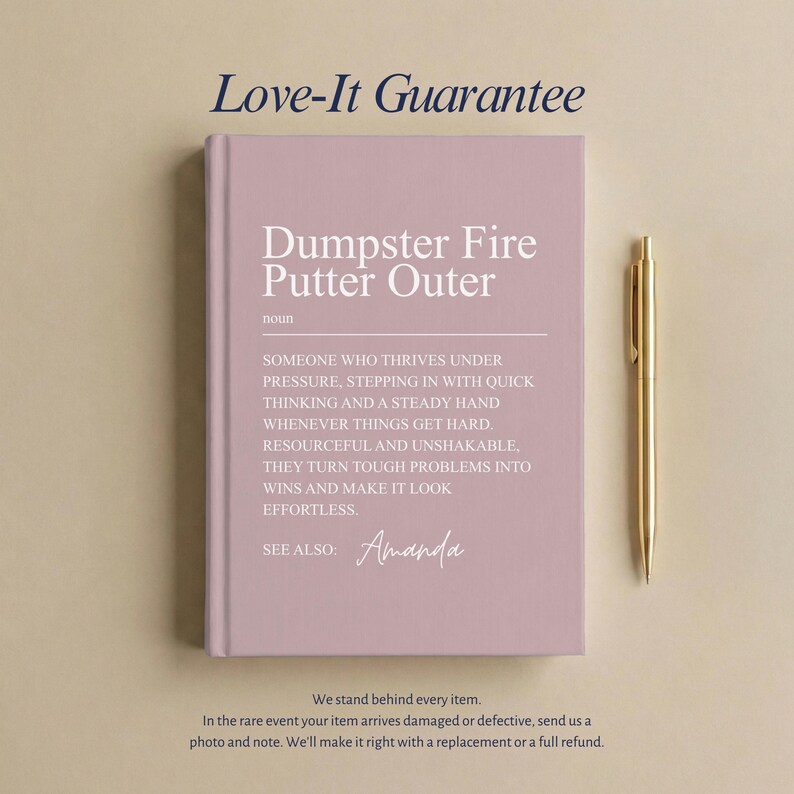 May include: A dusty rose hardcover journal featuring the white printed text Dumpster Fire Putter Outer, a definition of the term, and the name Amanda, accompanied by a gold-toned metal pen positioned vertically to the right of the book.
