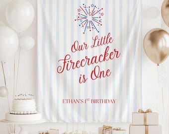 May include: A festive birthday banner with blue and white stripes, featuring red text that reads "Our Little Firecracker is One." The banner also includes the text "Ethan's 1st Birthday" and a firework graphic. White and gold balloons and gifts are also present.
