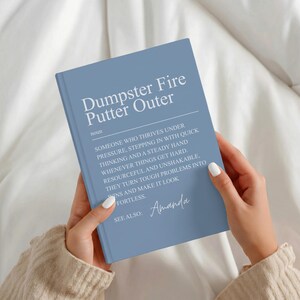 May include: A dusty blue hardcover book with white text on the front cover. The text reads: Dumpster Fire Putter Outer, noun, someone who thrives under pressure, stepping in with quick thinking and a steady hand whenever things get hard. Resourceful and unshakable, they turn tough problems into wins and make it look effortless. See also: Amanda.