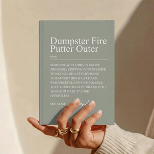 May include: A sage green hardcover book with white text on the front cover. The text reads: Dumpster Fire Putter Outer, noun, followed by a definition describing someone who thrives under pressure, and the phrase SEE ALSO: and.