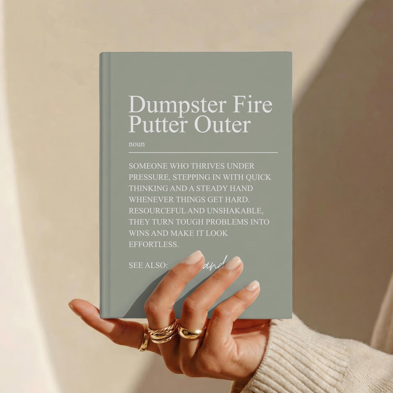 May include: A sage green hardcover book with white text on the front cover. The text reads: Dumpster Fire Putter Outer, noun, followed by a definition describing someone who thrives under pressure, and the phrase SEE ALSO: and.
