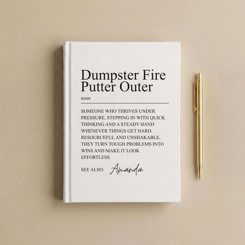 May include: A white hardcover journal with black text that reads Dumpster Fire Putter Outer, noun, followed by a definition describing someone who thrives under pressure, and the text See Also: Amanda, accompanied by a gold metal ballpoint pen.