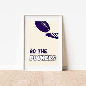 Puede incluir: Un póster enmarcado en blanco con un fondo crema que presenta un balón de fútbol morado que es pateado por un zapato morado. El texto "GO THE DOCKERS" está en una fuente morada desgastada.