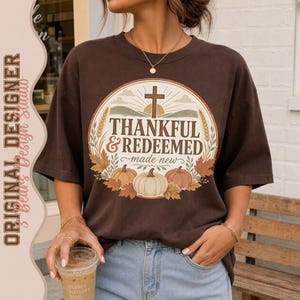 May include: A dark brown short-sleeve t-shirt featuring a circular graphic with a cross, sun rays, wheat stalks, and pumpkins. The text reads Thankful & Redeemed made new. The design includes autumn leaves in shades of orange, brown, and cream.