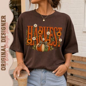 Könnte beinhalten: Ein dunkelbraunes Kurzarm-T-Shirt mit dem Wort HARVEST in großen orangefarbenen Buchstaben. Das Design zeigt gelbe Sonnenblumen, weiße Gänseblümchen und einen Kürbis mit einer schwarz-weiß karierten Schleife. Das Shirt hat einen Rundhalsausschnitt.