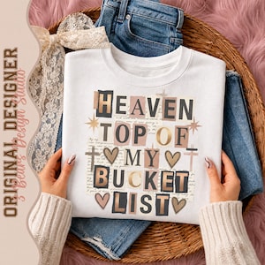 Könnte beinhalten: Ein weißes Sweatshirt mit Rundhalsausschnitt und einem Collage-Grafikdruck mit dem Text HEAVEN TOP OF MY BUCKET LIST. Das Design enthält schwarze und goldene Buchstaben, kleine Herzsymbole, Kreuze und Sterne auf einem handschriftlichen Hintergrund.