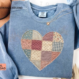 Puede incluir: Sudadera azul claro con un diseño de corazón de retazos. El corazón está hecho de varios patrones de tela en tonos rosa, azul y crema. Se ve un collar de oro con un colgante.