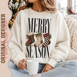 May include: A cream-colored crewneck sweatshirt featuring the black text MERRY SEASON and three illustrated Christmas stockings. The stockings display a red and green plaid pattern, a cream floral print, and a black and white checkered design.