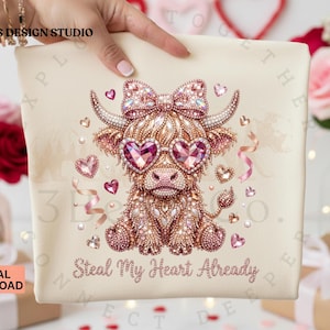 May include: Cream-colored t-shirt featuring a rhinestone-studded Highland cow wearing heart-shaped sunglasses and a bow. The text "Steal My Heart Already" is below the cow. Pink hearts and ribbons decorate the design.