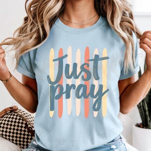 Może przedstawiać: Jasnoniebieska koszulka z napisem "Just Pray" w ozdobnej czcionce. Tekst jest nałożony na pionowe paski w odcieniach żółtego, pomarańczowego i niebieskiego. Koszulka ma krótkie rękawy i okrągły dekolt.