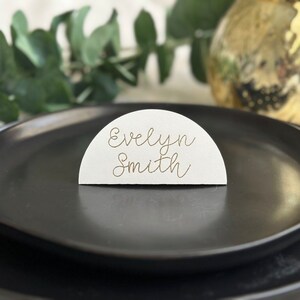 May include: White half-circle place card with the name "Evelyn Smith" written in gold calligraphy on a black plate.