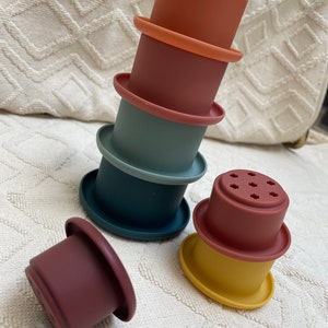 May include: A set of six colourful silicone stacking cups, featuring a variety of colours including red, orange, yellow, green, and blue. The cups are designed for bath time or sensory play.