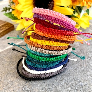 Può includere: Una pila di braccialetti in macramè colorati in varie tonalità, tra cui rosa, rosso, arancione, giallo, verde, blu e grigio. Ogni braccialetto ha un design annodato e lacci regolabili. Un bouquet di girasoli è sullo sfondo.