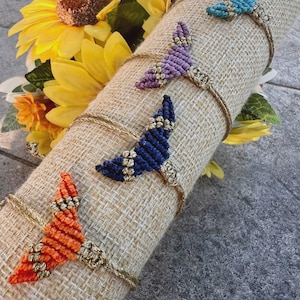 Peut inclure: Quatre bracelets en macramé en forme de queue de sirène, orange, violet, bleu et turquoise. Chaque queue est attachée à une chaîne dorée. Les bracelets sont présentés sur un tissu de jute roulé, avec des tournesols jaunes en arrière-plan.