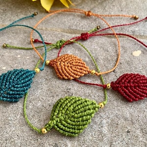 Könnte beinhalten: Makramee-Armbänder mit Blättern in Türkis, Orange, Rot und Grün. Jedes Armband hat ein gewebtes Blattdesign mit goldfarbenen Perlen und einem verstellbaren Kordelverschluss. Die Armbänder werden auf einer strukturierten grauen Oberfläche präsentiert.