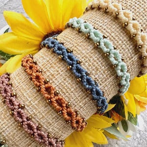 Peut inclure: Plusieurs bracelets en macramé de différentes couleurs : vert, rose, orange, bleu et crème. Chaque bracelet présente un motif tissé avec de petites perles dorées. Les bracelets sont présentés sur un cylindre enveloppé de toile de jute, avec des tournesols jaunes en arrière-plan.