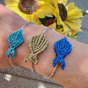 Könnte beinhalten: Drei Makramee-Armbänder in Türkis, Gold und Blau werden gezeigt. Jedes Armband hat ein geknotetes Design und ist an einer dünnen, neutralfarbenen Kordel befestigt. Die Armbänder werden vor einem Hintergrund aus gelben Sonnenblumen gezeigt.