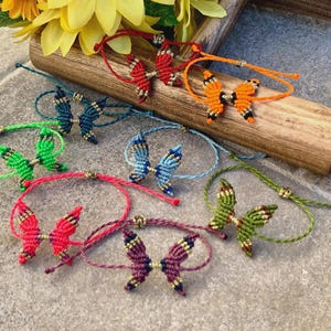 Puede incluir: Una colección de pulseras de mariposa hechas a mano en varios colores, incluyendo rojo, naranja, verde y azul. Cada pulsera presenta un diseño de mariposa de macramé y cordón ajustable. Las pulseras se exhiben sobre una superficie de madera.