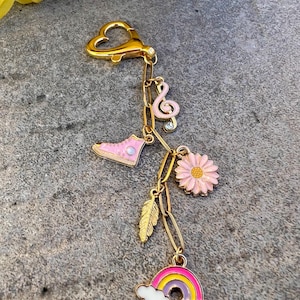 Può includere: Un portachiavi color oro con chiusura a forma di cuore e ciondoli multipli. I ciondoli includono una scarpa da ginnastica rosa, una nota musicale, una margherita, una piuma e un arcobaleno. I ciondoli sono collegati da catene color oro.