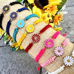 Può includere: Una collezione di braccialetti con ciondoli a forma di margherita in vari colori, tra cui nero, blu, turchese, giallo, rosso, rosa e bianco. Ogni braccialetto presenta un cordino intrecciato e una chiusura regolabile. I ciondoli a margherita hanno bordi in metallo dorato.