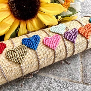 Könnte beinhalten: Eine Sammlung handgefertigter Makramee-Herzarmbänder in verschiedenen Farben, darunter Rot, Gold, Blau, Pink, Lila, Orange und Türkis. Die Armbänder werden auf einer strukturierten, beigefarbenen Stoffrolle mit einer Sonnenblume im Hintergrund präsentiert.