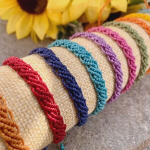 Könnte beinhalten: Eine Sammlung farbenfroher Makramee-Armbänder in Orange, Rot, Blau, Türkis, Lila, Rosa, Grün und Gelb. Die Armbänder werden auf einer beigen Stoffrolle präsentiert, mit einer gelben Sonnenblume im Hintergrund.