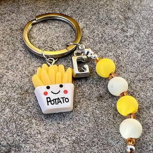 Può includere: Un portachiavi argentato con un ciondolo a forma di contenitore di patatine fritte. Il contenitore è bianco con la scritta "POTATO" e una faccina sorridente. Il portachiavi include anche un ciondolo con la lettera "G" argentata e un accento di perline gialle e bianche.