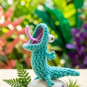 May include: A turquoise crochet alligator with a pink tongue and purple mouth sits on a gray stone. The handmade reptile has a white and brown eye. Ferns and a purple flower with green accents surround the stone, set against a blurred green background.