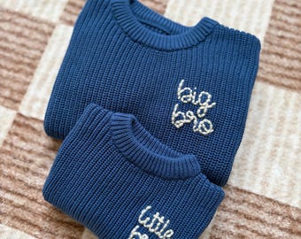May include: Two navy blue knit sweaters, one larger and one smaller, are stacked on top of each other. The larger sweater has "big bro" embroidered in white, and the smaller sweater has "little bro" embroidered in white.