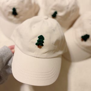 May include: Off-white baseball cap with a dark green embroidered tree design on the front. The cap has a curved brim and a fabric strap closure. Several similar caps are visible in the background.