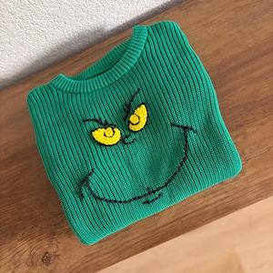 May include: A bright green ribbed knit sweater featuring a hand-stitched graphic of a face with two yellow eyes and a wide black embroidered smile. The crew neck pullover is made of a thick, textured yarn and displays a mischievous expression.
