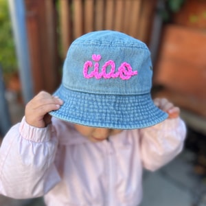 May include: A blue denim bucket hat with the word "ciao" embroidered in bright pink thread. The hat is being held by a person wearing a pink jacket.