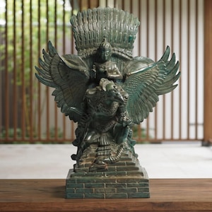 May include: A dark green bronze-toned sculpture depicting a figure mounted on a large winged creature with expansive feathers. The figure sits atop a tiered base, featuring intricate carvings and a weathered metallic finish throughout the entire piece.