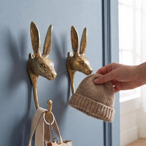 Puede incluir: Dos ganchos de cabeza de conejo de latón montados en una puerta azul. Se sostiene un gorro de punto beige cerca de los ganchos. Una bolsa de mano a rayas y un llavero de animal de madera cuelgan del gancho inferior. Decoración del hogar.