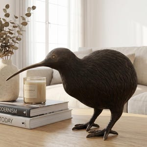 Op de afbeelding: Een donkerbruin kiwi vogelbeeld staat op een licht houten salontafel. Daarnaast liggen boeken met de titels "American Modernism" en "Sculpture Today", een vaas met gedroogde takken en een kaars. De kamer heeft een neutraal kleurenpalet.